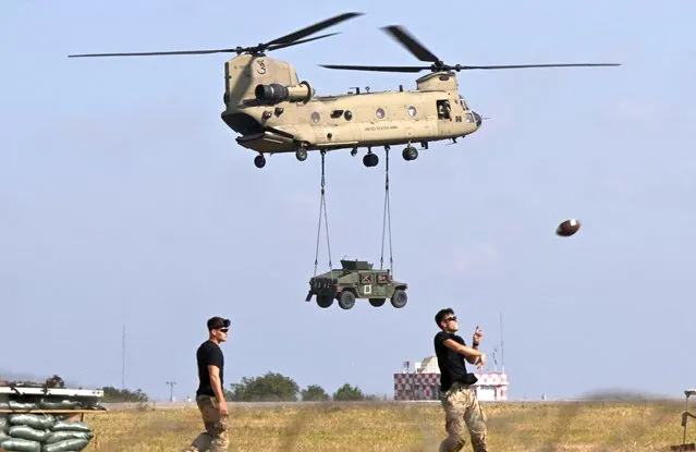 A Boeing CH-47F Chinook tandem rotor helicopter (Vertol) of the US Army's 101st Airborne Division transports a military vehicle while in the foreground military personnel play American football during a demonstration drill at Mihail Kogalniceanu Airbase near Constanta, Romania on July 30, 2022. Members of the 101st Airborne Division (Air Assault) headquarters and its 2nd Brigade Combat Team in a ceremony marked their official arrival in Europe at the airbase. The ceremony was followed by a “Romania/US Air and Land Showcase”, a combined US Army and Romanian Army capabilities demonstration. (Photo by Daniel Mihailescu/AFP Photo)