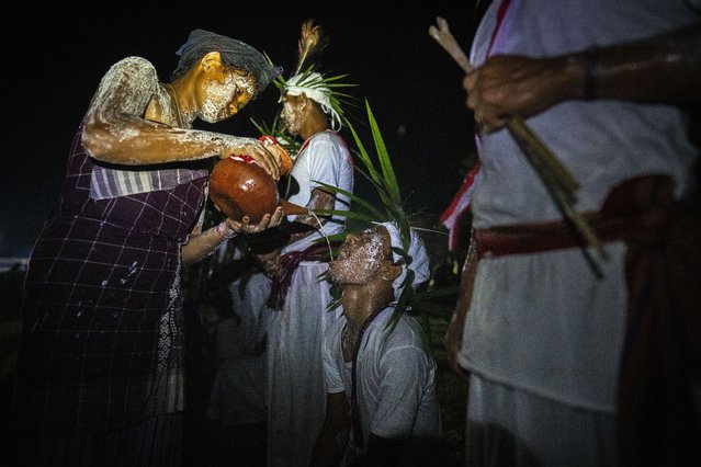 An Indian Rabha tribal girl in traditional attire with face smeared with rice flour pours rice beer as holy water to a Hindu priest after he walks barefoot over burning charcoal as part of rituals during Baikho festival at Gamerimura village along the Assam Meghalaya border, west of Gauhati, India, Saturday, June 4, 2022. (Photo by Anupam Nath/AP Photo)