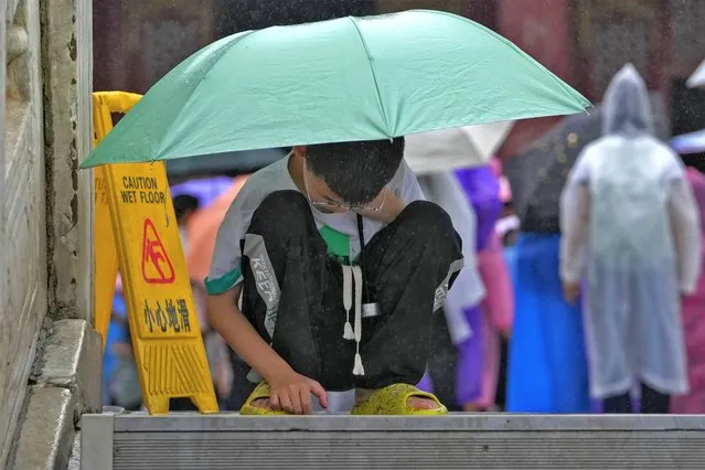 A boy covering himself with an umbrella from the rain, browses a smartphone placed on the ground at the Forbidden City in Beijing on July 13, 2023. China's internet watchdog has laid out regulations to curb the amount of time children spend on their smartphones, in the latest blow to firms such as Tencent and ByteDance, which run social media platforms and online games. (Photo by Andy Wong/AP Photo)