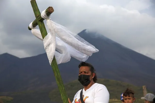 A woman carries a wooden cross during a pilgrimage with images of the town's patron saint San Vicente and the Virgin Mary, to pray for that the Pacaya Volcano decreases its activity, in San Vicente Pacaya, Guatemala, Wednesday, April 21, 2021. Residents of small communities living around Pacaya volcano wake up each morning wondering if the lava from the current eruption will reach their homes. (Photo by Moises Castillo/AP Photo)
