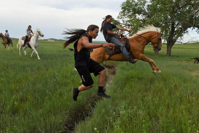 Mahto In The Woods jumps over a small creek on foot while his cousin Jayden Lookinghorse jumps over on his horse on the Cheyenne River Reservation in Green Grass, South Dakota, May 31, 2018. (Photo by Stephanie Keith/Reuters)