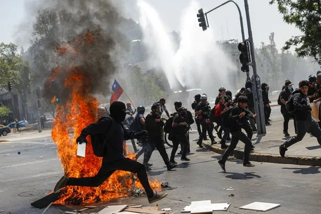Demonstrators run from police water cannons on the three-year anniversary of the start of anti-government protests against inequality, in Santiago, Chile, Tuesday, October 18, 2022. Protests that were sparked by a subway fare hike broke out on Oct. 18, 2019 and quickly broadened into a social movement demanding greater equality. (Photo by Cristobal Escobar/AP Photo)