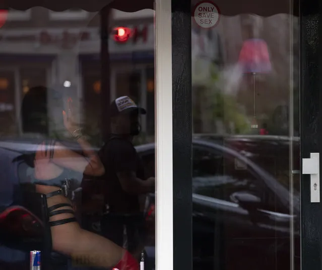 A sеx worker shields her face from the photographer as a passing man is reflected in the window after a further ease of corona virus restrictions in Amsterdam, Netherlands, Wednesday, July 1, 2020. It wasn't quite business as usual as the capital's Red Light District emerged from coronavirus lockdown, but it was as close as it has been since the pandemic slammed the brakes on the world's oldest profession. (Photo by Peter Dejong/AP Photo)