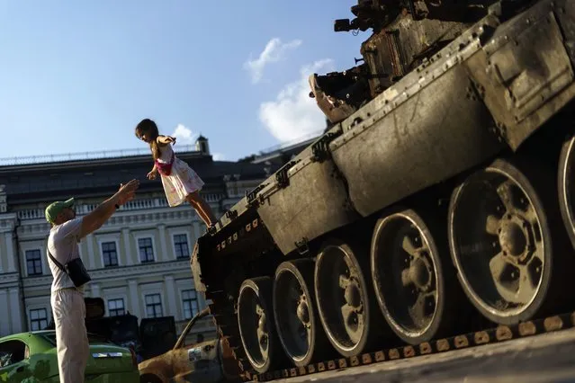 A young girl falls into her father's arms off a Russian tank that was destroyed and exhibited at Mykhailivs'ka Square with other damaged Russian military equipment as a symbol of Ukraine's resistance against the invasion in Kyiv, Ukraine, Saturday, July 30, 2022. (Photo by David Goldman/AP Photo)