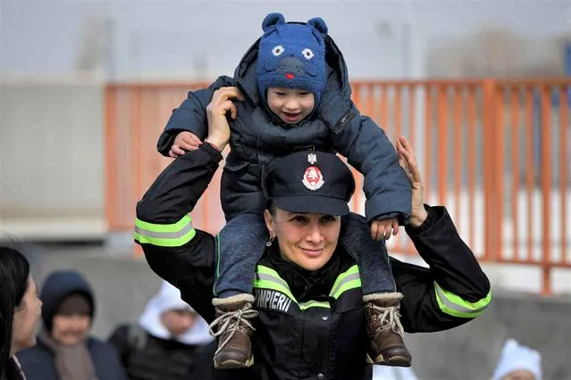 A firefighter holds a child on her shoulders after his family fleeing the war from neighbouring Ukraine crossed the border by ferry at the Isaccea-Orlivka border crossing in Romania, Friday, March 25, 2022. In Ukrainian towns and cities that day-by-day increasingly resemble the ruins that Russian forces left behind in previous campaigns in Syria and Chechnya, the misery for civilians grows ever more acute. (Photo by Andreea Alexandru/AP Photo)