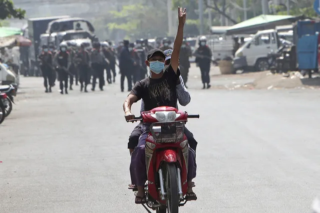 A protester on a motorbike flashes the three-fingered salute while soldiers and police take position to disperse protesters on one of the main roads in Mandalay, Myanmar, Friday, February 26, 2021. Security forces in Myanmar's largest city Yangon on Friday fired warning shots and beat truncheons against their shields while moving to disperse more than 1,000 anti-coup protesters. (Photo by AP Photo/Stringer)
