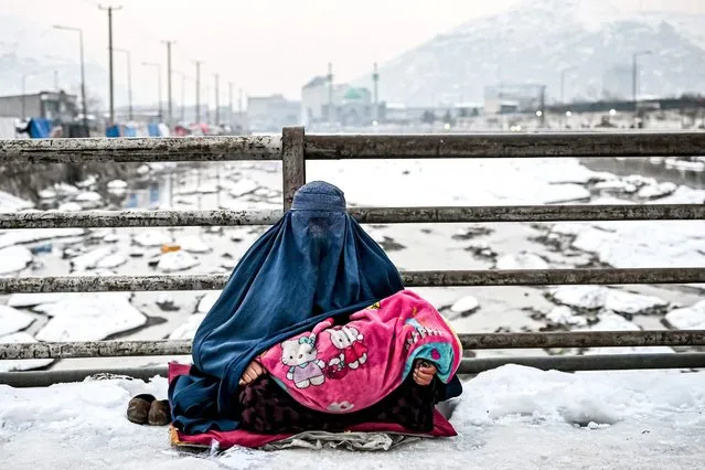 A burqa-clad Afghan woman sits with a child on her lap as she seeks alms from passers-by on a bridge covered with snow in Kabul on January 6, 2022. (Photo by Mohd Rasfan/AFP Photo)