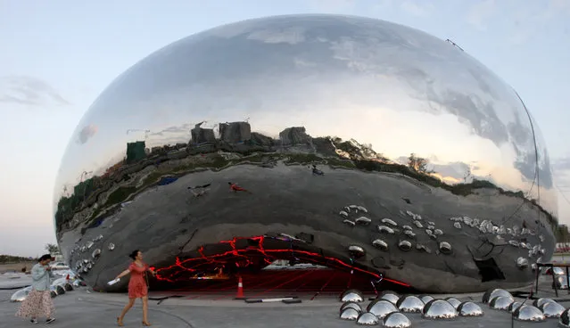 In this photo taken August 9, 2015, residents walk past a giant sculpture in the shape of an oil bubble under construction in the city of Karamay in the China's Xinjiang Uighur Autonomous Region. Renowned British-Indian artist Anish Kapoor has expressed outrage about the appearance of the sculpture in China that appears identical to his “Cloud Gate” in Chicago. (Photo by Color China Photo via AP Photo)
