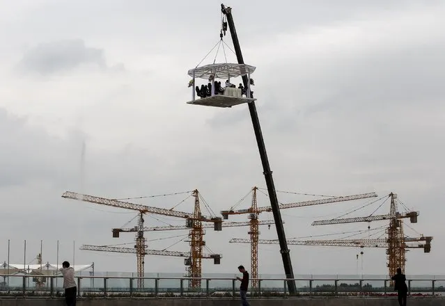 Bystanders look on as people dine at a table lifted by a crane to a height of 30 metres mid-air, near a residential construction site, in Kunming, Yunnan province, China, July 23, 2015. According to local media, having a meal at this lifted dining table can cost 8,888 yuan (1,431 USD) for one person. (Photo by Wong Campion/Reuters)