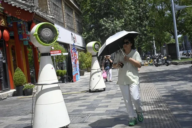 A woman using an umbrella to shelter from the sun walks past a cooling fan during hot weather conditions in Beijing on July 6, 2023. (Photo by Jade Gao/AFP Photo)