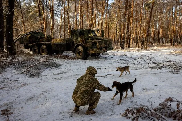 A serviceman of Ukraine's 45th separate artillery brigade feeds a stray dog near a Swedish-made Archer self-propelled howitzer system in a forest in the Donetsk region, amid Russia’s attack on Ukraine on December 16, 2023. (Photo by Thomas Peter/Reuters)