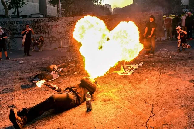 A protester spits fire during a protest to mark the second anniversary of months of civil unrising against social inequality, in Santiago, on October 18, 2021. The social-justice protests that rocked Chile in October 2019 led ultimately to the decision to create a new constitutional convention to draft a constitution to replace the one written during the dictatorship of Augusto Pinochet (1973-1990). The new 155-member the new body is in charge of writing a new constitution meant to pry power from the hands of the elite and spread it more equitably in the South American nation. (Photo by Martin Bernetti/AFP Photo)