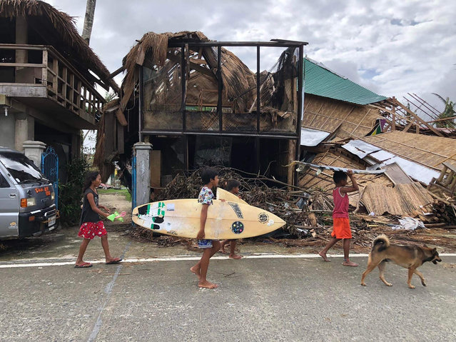 In this photo taken on December 29, 2021, young residents carrying their surf boards, walk past houses destroyed by super Typhoon Rai, as they head for the beach in General Luna town, Siargao island, more than a week after the super typhoon devastated the island. (Photo by Roel Catoto/AFP Photo)