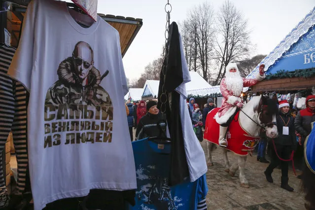 An actors dressed as Grandfather Frost, Russian Santa Claus, rides past a kiosk selling T-shirts with a portrait of the Russian President Vladimir Putin during celebrations for the upcoming Orthodox Christmas at a Christmas market in St.Petersburg, Russia, Wednesday, January 6, 2016. The sign on the T-shirt signs 'The most polite man'. (Photo by Dmitry Lovetsky/AP Photo)