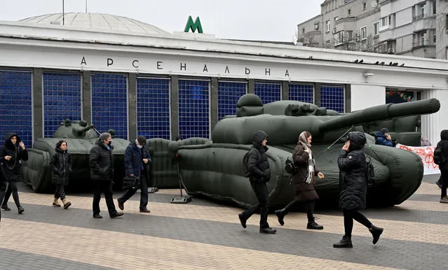 People walk past inflatable tanks placed outside a metro station in the Ukrainian capital of Kiev on December 16, 2021, during a protest action held by workers of a small private company producing inflatable dummies of military equipment for the armed forces after the Defence Ministry refused to pay for its order. (Photo by Sergei Supinsky/AFP Photo)