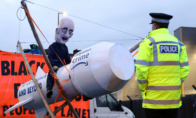 A police officer looks at a person wearing a head mask depicting Amazon founder, Jeff Bezos, as Extinction Rebellion activists block an entrance to an Amazon fulfilment centre in Tilbury, Essex, Britain, November 26, 2021. (Photo by Henry Nicholls/Reuters)