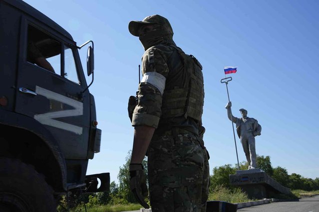 A military truck with the letter Z, which has become a symbol of the Russian military, drives past a Russian soldier standing in the road at the entrance of Mariupol with a Soviet style symbolic monument of a metallurgist, on the territory which is under the Government of the Donetsk People's Republic control, eastern Ukraine, Sunday, June 12, 2022. This photo was taken during a trip organized by the Russian Ministry of Defense. (Photo by AP Photo/Stringer)