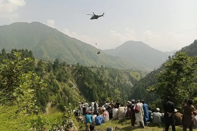 People watch as an army soldier slings down from a helicopter during a rescue mission to recover students stuck in a chairlift in Pashto village of mountainous Khyber Pakhtunkhwa province, on August 22, 2023. Six children and two adults were suspended inside a cable car dangling over a deep valley in Pakistan for several hours on August 22, as a military helicopter hovered nearby. (Photo by Prateek Kumar/AFP Photo)