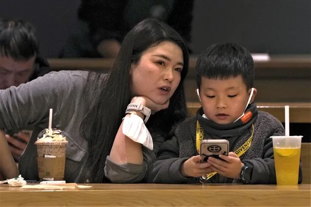 A woman talks to a child playing with his smartphone at a cafe in Beijing, Thursday, January 19, 2023. As the week-long Lunar New Year holidays in China draw near with promises of feasts and red envelopes stuffed with cash, children have yet another thing to look forward to – one more hour of online games each day. (Photo by Andy Wong/AP Photo)