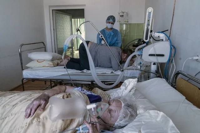 An elderly woman suffering from COVID-19 breathes with the help of an oxygen mask, foreground, as a nurse treats a patient in central district hospital of Kolomyia, western Ukraine, Tuesday, February 23, 2021. After several delays, Ukraine finally received 500,000 doses of the AstraZeneca vaccine marketed under the name CoviShield, the first shipment of Covid-19 vaccine doses. The country of 40 million is one of the last in the region to begin inoculating its population. (Photo by Evgeniy Maloletka/AP Photo)