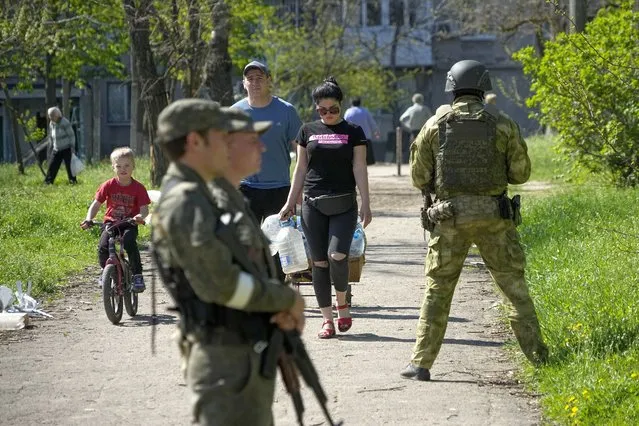 Serviceman of Donetsk People's Republic militia guard an area as local civilians walk to get humanitarian aid, bread and pure water distributed by Donetsk People Republic Emergency Situations Ministry in Mariupol, in territory under the government of the Donetsk People's Republic, eastern Ukraine, Friday, April 29, 2022. This photo was taken during a trip organized by the Russian Ministry of Defense. (Photo by AP Photo/Stringer)