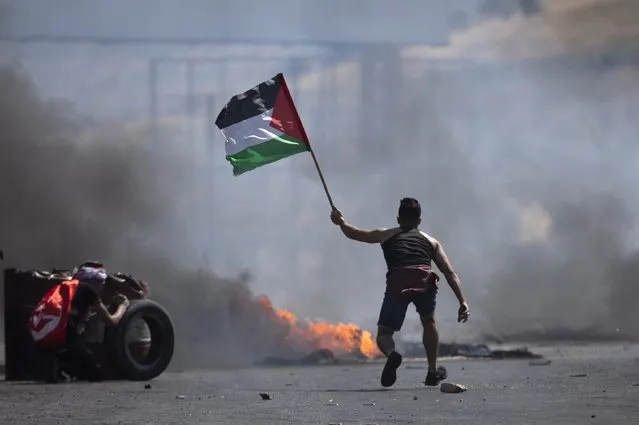 A protester waves the Palestinian flag during clashes with the Israeli forces at the Hawara checkpoint, south of the West Bank city of Nablus, Friday, May 14, 2021. Health officials say several Palestinians were killed by Israeli army fire, at protests that took place in several locations across the West Bank of Friday. (Photo by Majdi Mohammed/AP Photo)