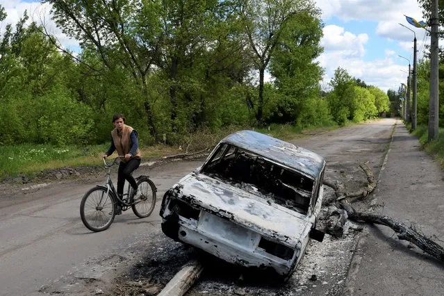A man rides a bicycle past a car destroyed by shelling in a street in the village of Niu-York, Donetsk region, Ukraine, Monday, May 16, 2022. (Photo by Andriy Andriyenko/AP Photo)