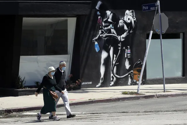 People wearing masks walk past a mural depicting a coronavirus theme during the COVID-19 pandemic Tuesday, April 14, 2020, in Los Angeles. California Gov. Gavin Newsom on Tuesday unveiled an outline for what it will take to lift coronavirus restrictions in the nation's most populous state, asking more questions than answering them as he seeks to temper the expectations of a restless, isolating public. (Photo by Marcio Jose Sanchez/AP Photo)