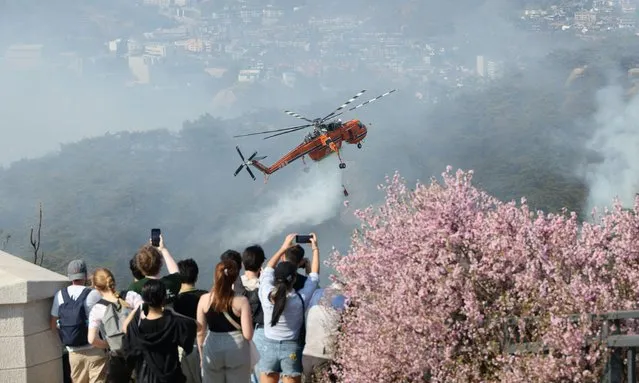 This picture taken on April 2, 2023 shows people watching as a helicopter douses a wildfire on Mount Inwang in Seoul. (Photo by Yonhap via AFP Photo)