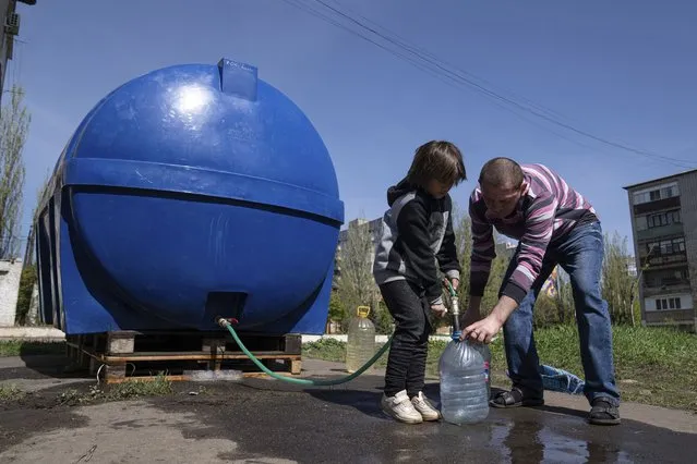 A girl Lisa helps a man to fill cans with water from a water tank installed for residents of Toretsk, eastern Ukraine, Monday, April 25, 2022. Toretsk residents have had no access to water for more than two months because of the war. (Photo by Evgeniy Maloletka/AP Photo)