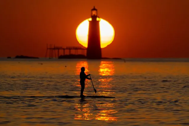 Paula Birmingham of South Portland, Maine, glides along on her stand up paddle board as the sun rises behind Ram Island Ledge Light, Thursday morning, August 24, 2023, off Cape Elizabeth, Maine. (Photo by Robert F. Bukaty/AP Photo)