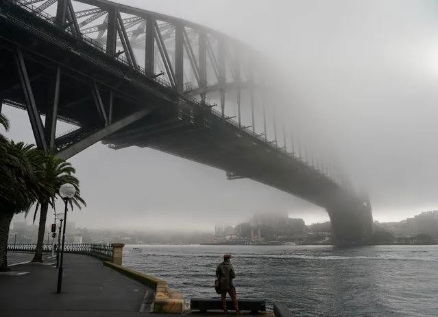 A man wearing a protective face mask takes in the waterfront view underneath the Sydney Harbour Bridge, seen shrouded in fog, during a lockdown to curb the spread of a coronavirus disease (COVID-19) outbreak in Sydney, Australia, June 30, 2021. (Photo by Loren Elliott/Reuters)