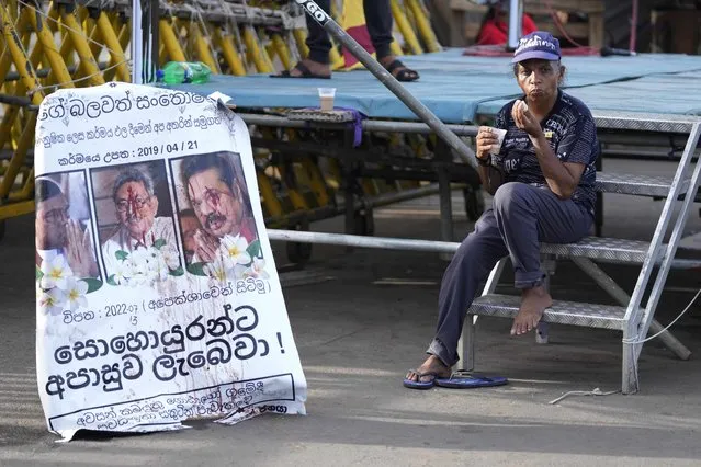 A protester drinks a cup of tea as she sits by a defaced poster carrying portraits of ousted president Gotabaya Rajapaksa, center, and his brothers at the entrance to president's office in Colombo, Sri Lanka, Friday, July 15, 2022. Protesters retreated from government buildings Thursday in Sri Lanka, restoring a tenuous calm to the economically crippled country, and the embattled president at last emailed the resignation that demonstrators have sought for months. (Photo by Eranga Jayawardena/AP Photo)