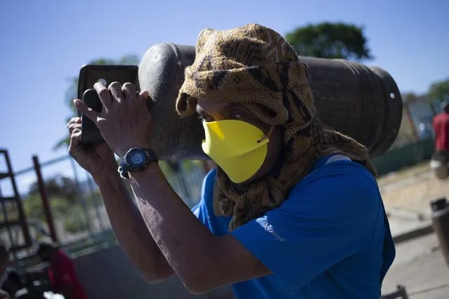 A man wearing homemade face protection carries a full cylinder of cooking gas after waiting in a long line for the gas truck to arrive during a lockdown to contain the spread of the new coronavirus in Caracas, Venezuela, Friday, March 27, 2020. (Photo by Ariana Cubillos/AP Photo)
