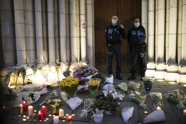 Police stand next to flowers and candles set on the steps of the Notre Dame church in Nice, France, Friday, October 30, 2020. A new suspect is in custody in the investigation into a gruesome attack by a Tunisian man who killed three people in a French church. France heightened its security alert amid religious and geopolitical tensions around cartoons mocking the Muslim prophet. (Photo by Daniel Cole/AP Photo)