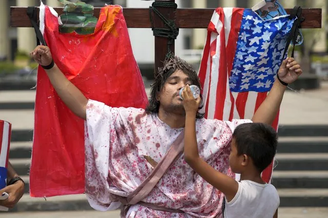 A child gives water to an activist strapped on a wooden cross with the mock flag of the United States and China as they reenact the sufferings of Jesus Christ during a holy week protest in Manila, Philippines on Monday April 3, 2023. The group gathered to raise awareness of the hardships people are suffering as Holy Week begins in this predominantly Roman Catholic nation. (Photo by Aaron Favila/AP Photo)