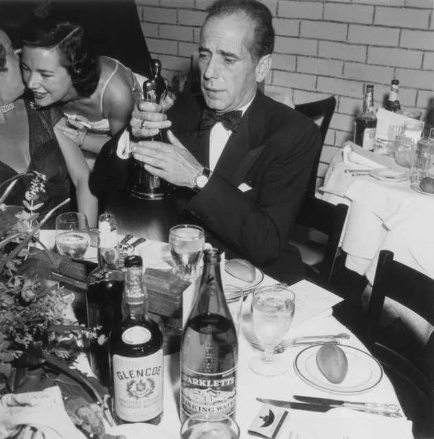 American actor Humphrey Bogart (1899 – 1957) sits at a table with place settings and liquor bottles, holding up his Best Actor statue at the Academy Awards, RKO Pantages Theater, Los Angeles, California on March 20, 1952. Bogart won the Oscar for his role in director John Huston's film, “The African Queen”. (Photo by Murray Garrett/Getty Images)