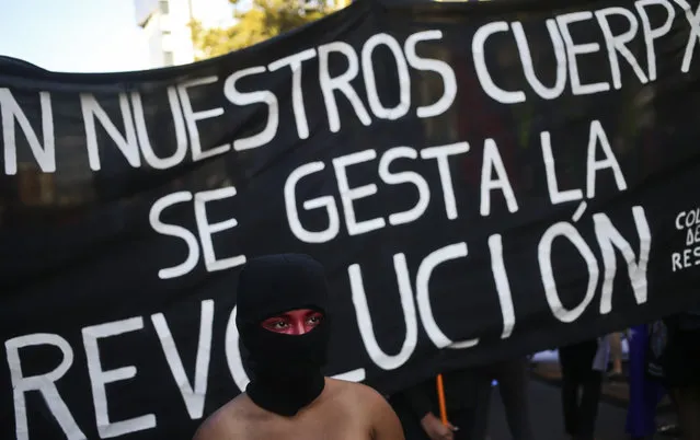 A masked woman walks in front of a banner that says in Spanish: “The revolution gestates in our bodies” at a march marking International Women's Day outside La Moneda presidential palace in Santiago, Chile, Wednesday, March 8, 2017. Many women stayed home from work, joined rallies or wore red Wednesday as International Women's Day was observed with a multitude of events around the world. (Photo by Esteban Felix/AP Photo)