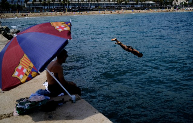 A man jumps into the Mediterranean Sea as his friend sits under an umbrella with an FC Barcelona theme, at Barceloneta beach in Barcelona, Spain on August 25, 2024. (Photo by Nacho Doce/Reuters)