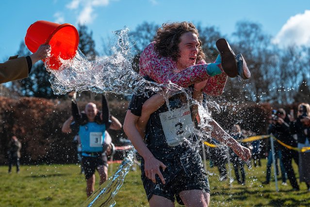 Competitors have water thrown over them as they take part in the annual UK wife carrying race on March 16, 2025 in Dorking, England. The UK Wife Carrying Race was first held in 2008, and features either males or females carrying a 'wife' who can be anyone over 18 that meets the weigh-in requirements. Competitors traverse the 380m course up and down The Nower, a steep hill with a 15m ascent. (Photo by Carl Court/Getty Images)