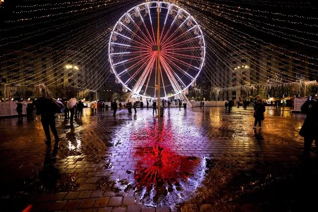 An illuminated Ferris wheel is reflected in the wet pavement at a Christmas fair which opened ahead of the holiday season in Bucharest, Romania, November 20, 2022. In cities across Europe, officials are wrestling with a choice this Christmas. Dim lighting plans to send a message of energy conservation and solidarity with citizens squeezed by both higher energy costs and inflation or let the lights blaze in a message of defiance after two years of pandemic-suppressed Christmas seasons, creating a mood that retailers hope loosen holiday purses. (Photo by Andreea Alexandru/AP Photo)