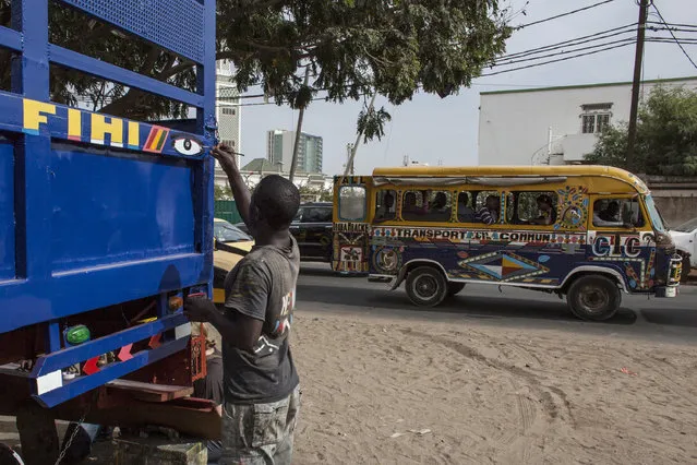 In this photo taken Saturday, December 19, 2015, Kalidou Diallo, known as “Neyoo”, paints the side panel of a car rapide, normally used for transporting goods and luggage, as the version of the “car rapide” that will be phased out as of next year passes behind him in Dakar, Senegal. People like Diallo have made their distinctive mark on these vehicles that are moving art, whizzing through the streets of Dakar, people often hanging off the sides. (Photo by Jane Hahn/AP Photo)