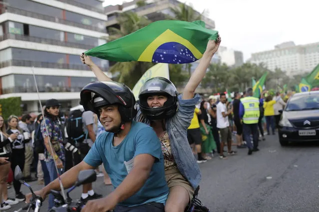 A supporter of presidential front-runner Jair Bolsonaro rides on the back of a motorcycle waving a national flag, outside Bolsonaro's residence in Rio de Janeiro, Brazil, Sunday, October 28, 2018. Bolsonaro took a commanding lead in the race for Brazil's presidency, as voters apparently looked past warnings that the brash former army captain would erode democracy and embraced a chance for radical change after years of turmoil. (Photo by Silvia Izquierdo/AP Photo)