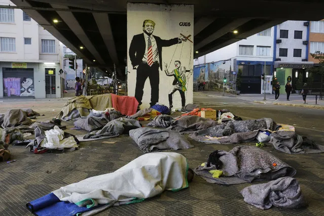 Homeless people sleep under a bridge in front of a mural depicting U.S. President Donald Trump as a puppeteer manipulating Brazil's President Jair Bolsonaro, in downtown Sao Paulo, Brazil, Tuesday, July 23, 2019.  Bolsonaro said last week that severe hunger is not a problem for the country. He acknowledged that things could “go bad” and people might “not eat well”. But, he added: “To speak of starving in Brazil is a big lie”. (Photo by Nelson Antoine/AP Photo)