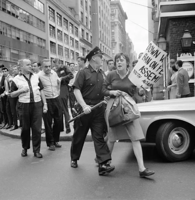 Woman demonstrator carrying a paper bag and a torn placard is escorted by New York City policeman in the area of Fifth Avenue and 47th street on August 15, 1964. She was one of 250 demonstrators who attempted to hold a rally in Duffy Square to protest U.S. action in Vietnam. They were battled by police and about thirty arrests made when they attempted to walk towards the United Nations headquarters. Rally was called by the “May 2 Movement” and “Youth against War and Fascism” organizations. (Photo by AP Photo)