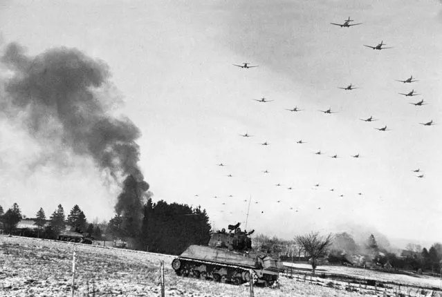 Low flying C-47 transport planes roar overhead as they carry supplies to the besieged American Forces battling the Germans at Bastogne, during the enemy breakthrough on January 6,1945 in Belgium. In the distance, smoke rises from wrecked German equipment, while in the foreground, American tanks move up to support the infantry in the fighting. (Photo by AP Photo)