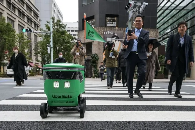 Members of the media and passersby walk next to an unmanned robot as it crosses the street during a demonstration of a robot delivery service by Uber Eats Japan, Mitsubishi Electric and robot developer Cartken in downtown Tokyo on March 5, 2024. (Photo by Richard A. Brooks/AFP Photo)