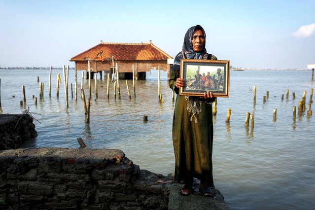 Pasijah, 55, carries a framed photograph of her family as she stands at her home, with a partially submerged building in the background, in the hamlet of Rejosari Senik, Demak regency, Central Java Province, Indonesia, March 14, 2025. Pasijah's home is the only one remaining in this part of Rejosari Senik, a small village on Java's northern coast that was once on dry land but is now submerged by water, as over the past few years, her neighbours have abandoned their homes, vegetable plots and rice fields to the advancing sea. “I do have every intention to stay here and my feelings for this house remain”, she told Reuters in February. (Photo by Ajeng Dinar Ulfiana/Reuters)