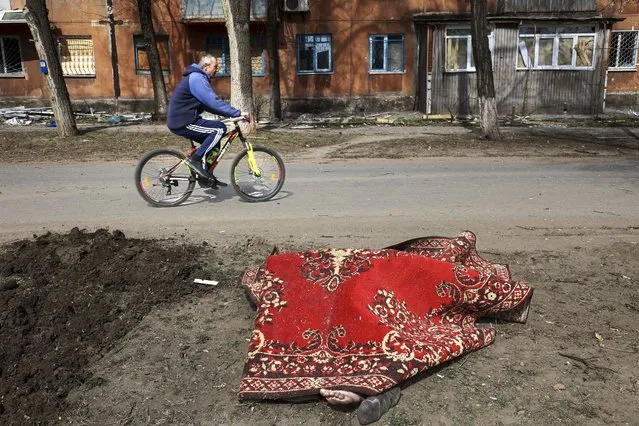 A man rides his bicycle past a body covered by a rug after fighting on the outskirts of Mariupol, Ukraine, in territory under control of the separatist government of the Donetsk People's Republic, on Tuesday, March 29, 2022. (Photo by Alexei Alexandrov/AP Photo)