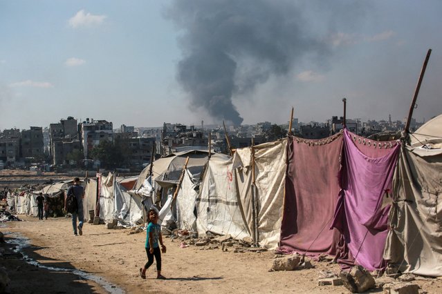 People walk past makesfift shelters as smoke billows east of Gaza City, in the central Gaza Strip during Israeli bombardment on July 6, 2025. An Israeli delegation was expected in Doha on July 6 for talks on a Gaza truce and hostage release deal, after Palestinian group Hamas said it was ready to start negotiations "immediately". (Photo by Bashar Taleb/AFP Photo)